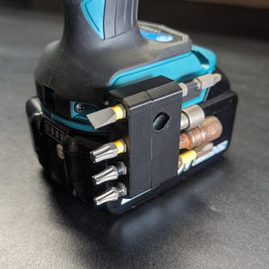 May include: A black 3D printed holder for drill bits attached to a teal and black drill battery. The holder has slots for six drill bits and a small hole for a screwdriver bit.