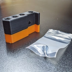 Puede incluir: Una pieza de pl&aacute;stico negra y naranja impresa en 3D con tres agujeros y un tornillo en una bolsa de pl&aacute;stico transparente.