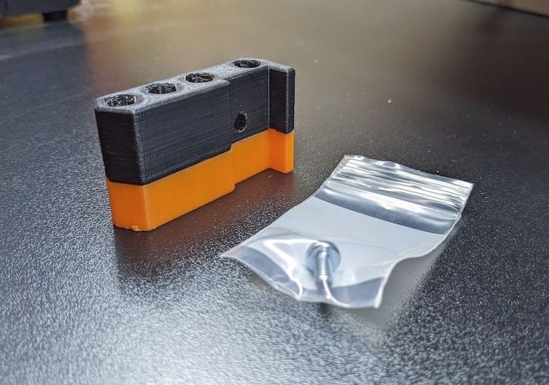 Puede incluir: Una pieza de pl&aacute;stico negra y naranja impresa en 3D con tres agujeros y un tornillo en una bolsa de pl&aacute;stico transparente.