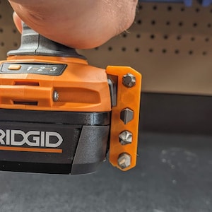 Puede incluir: Un taladro de bater&iacute;a naranja y negro con un logotipo RIDGID negro y plateado. El taladro tiene un soporte de brocas integrado con cinco brocas diferentes.
