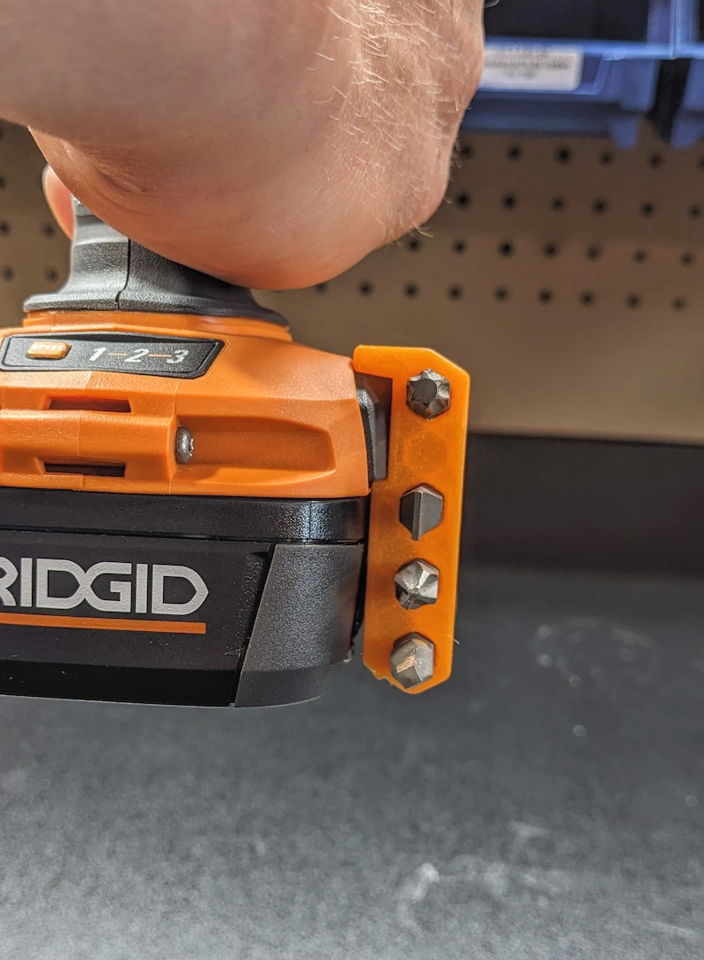 Puede incluir: Un taladro de bater&iacute;a naranja y negro con un logotipo RIDGID negro y plateado. El taladro tiene un soporte de brocas integrado con cinco brocas diferentes.