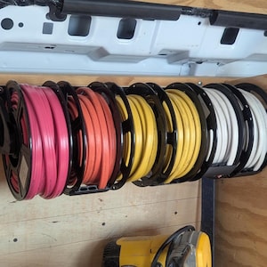 Pode incluir: Um suporte de armazenamento montado na parede com cinco carretéis circulares de plástico preto, cada um enrolado com uma mangueira ou cabo de cor diferente. As cores da esquerda para a direita são rosa, laranja, amarelo, amarelo e branco.