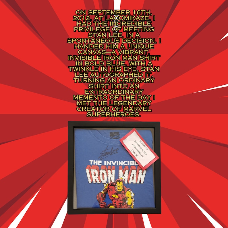 Op de afbeelding: Een ingelijst blauw T-shirt met een rode en gele Iron Man-afbeelding en de tekst "The Invincible Iron Man". Het shirt is gesigneerd door Stan Lee met de datum 16 september 2012.
