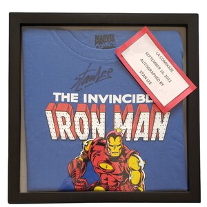 Op de afbeelding: Een ingelijst blauw T-shirt met een rode en gele Iron Man-afbeelding en de tekst "The Invincible Iron Man". Het shirt is gesigneerd door Stan Lee met een briefje waarop staat "La Comikaze September 16, 2012 Autographed by Stan Lee".