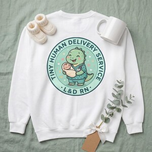 May include: A white crewneck sweatshirt featuring a circular graphic of a green dinosaur dressed as a nurse holding a swaddled baby. The text reads TINY HUMAN DELIVERY SERVICE and L&D RN. The design includes small hearts and stars on a light green background.
