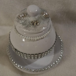 May include: A white ceramic trinket box with a lid, decorated with silver glitter, faux pearls, and a small silver elephant charm. The base has a beaded edge and a silver rhinestone trim. The lid has a white knob.