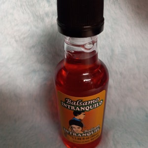 May include: A small glass bottle with a black cap filled with a red liquid. The label is yellow with black text that reads "Balsamo Intranquilo" and a black and white illustration of a couple. The label also says "Balsam Intranquil" and "1.69 fl oz (50 ml)".