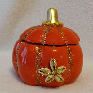 Puede incluir: Una caja de baratijas decorativa con forma de calabaza naranja con un tallo dorado y detalles de purpurina dorada. La caja presenta un diseño floral hecho de conchas marinas y purpurina dorada. La tapa es extraíble.