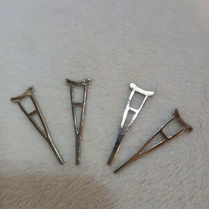 Puede incluir: Cuatro dijes plateados con forma de muleta se disponen sobre una superficie blanca y suave. Cada dije tiene una parte superior curvada, un centro rectangular y una base triangular, asemejándose a ayudas médicas en miniatura.