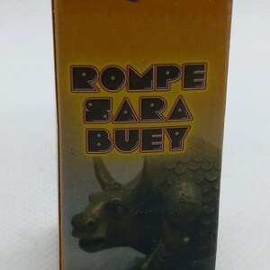 Peut inclure: Boîte rectangulaire jaune moutarde avec le mot "ACHE" en haut dans une police stylisée. En dessous, le texte "ROMPE SARA BUEY" est affiché en grandes lettres en gras. Une illustration de dinosaure est visible. Le bas de la boîte contient du texte supplémentaire.
