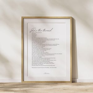 May include: A framed print with the text "She's the thread" in elegant script. The print features a poem about a woman's role in a family, with a light-colored wooden frame. The artwork is displayed on a light-colored surface.