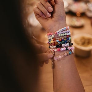May include: A person's wrist with a stack of colorful beaded bracelets. The bracelets have different colored beads and letters spelling out words like "OUR", "VIV", "SHA", "MOM", "IVY", "LOVER", and "KRAL".