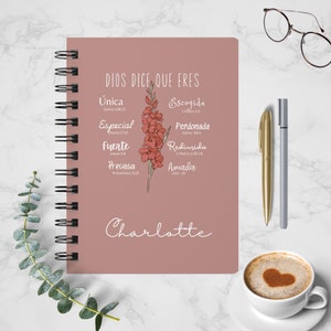 May include: A pink spiral bound journal with the text "DIOS DICE, QUE ERES" and a floral illustration. The journal has a list of words with bible verses, including "Unica", "Especial", "Fuerte", "Preciosa", "Escogida", "Perdonada", "Redimida", and "Amada". The name "Charlotte" is written at the bottom of the journal.