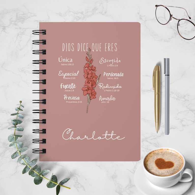 May include: A pink spiral bound journal with the text "DIOS DICE, QUE ERES" and a floral illustration. The journal has a list of words with bible verses, including "Unica", "Especial", "Fuerte", "Preciosa", "Escogida", "Perdonada", "Redimida", and "Amada". The name "Charlotte" is written at the bottom of the journal.