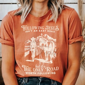 May include: A rust-colored t-shirt with a white graphic of a woman on horseback. The graphic includes the text "Following Jesus isn't an easy road, but it's the only road worth following."