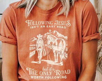 May include: A rust-colored t-shirt with a white graphic of a woman on horseback. The graphic includes the text "Following Jesus isn't an easy road, but it's the only road worth following."