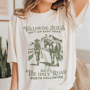 May include: A white t-shirt with a green graphic design of a woman on horseback. The text on the shirt reads "Following Jesus isn't an easy road, but it's the only road worth following."