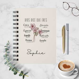 May include: A cream-colored notebook with a spiral binding features a floral design with pink flowers and green leaves. The notebook has the text "DIOS DICE QUE ERES" at the top and a list of words with corresponding Bible verses below. The name "Sophie" is written in cursive at the bottom of the notebook.