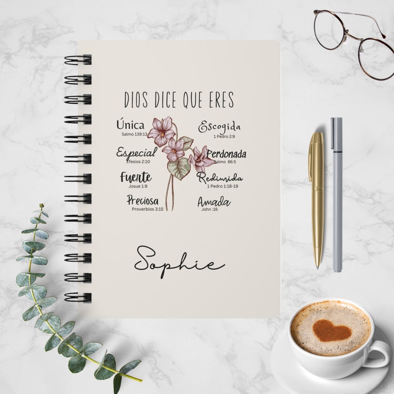 May include: A cream-colored notebook with a spiral binding features a floral design with pink flowers and green leaves. The notebook has the text "DIOS DICE QUE ERES" at the top and a list of words with corresponding Bible verses below. The name "Sophie" is written in cursive at the bottom of the notebook.
