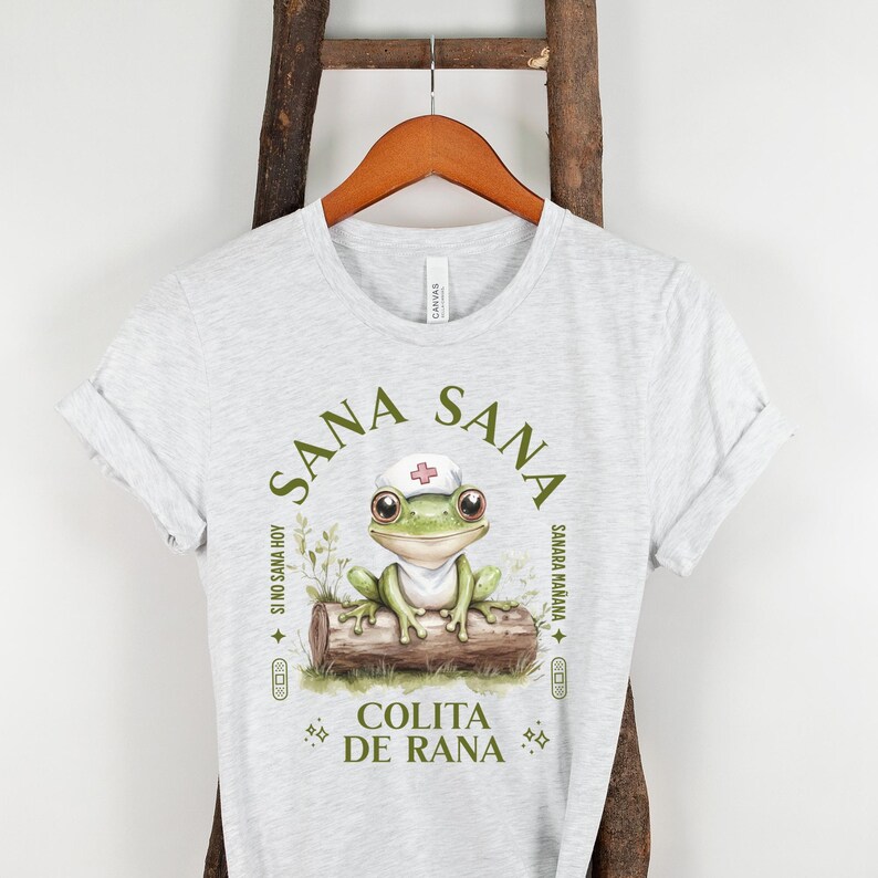May include: Gray t-shirt with a green frog wearing a white nurse's hat. The frog is sitting on a log with green leaves. The text on the shirt reads "Sana Sana Colita de Rana" with the words "Si no sana hoy, sanara ma&ntilde;ana" on the side.
