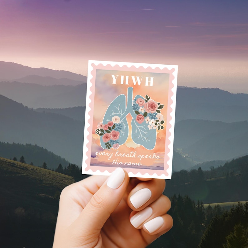 May include: A sticker with a light blue lung design adorned with floral arrangements. The sticker has the text "YWHW" at the top and "every breath speaks this name" at the bottom. The sticker is held in front of a scenic mountain backdrop.