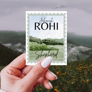 May include: A rectangular sticker with a watercolor landscape scene and the words "Jehovah Rohi" and "The Lord my Shepherd" in script. The sticker has a light green border and is held by a hand with white nail polish.