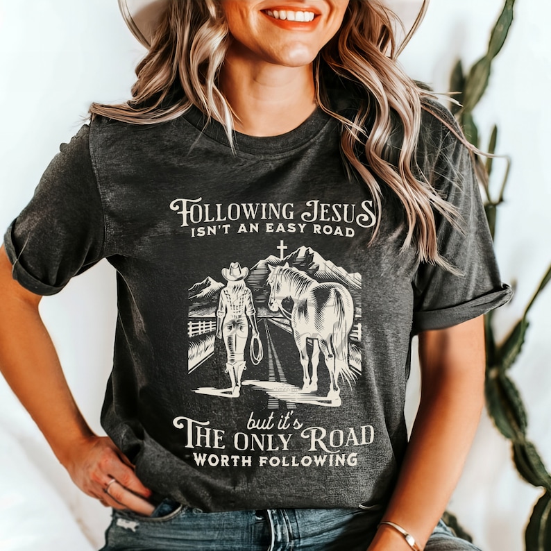 May include: A dark gray t-shirt with a graphic of a woman on horseback riding through a mountain landscape. The text on the shirt reads "Following Jesus isn't an easy road, but it's the only road worth following."