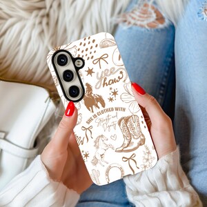 May include: White phone case with a brown and white western-themed design. The design includes a horse, cowboy boots, a horseshoe, a cactus, and the text "yee haw" and "She is clothed with strength and dignity".