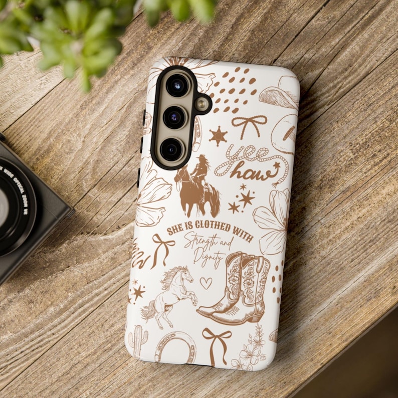 May include: White phone case with a brown and white design. The design includes a cowboy boot, a horse, a cactus, a horseshoe, a bow, and the text "Yee Haw" and "She is clothed with strength and dignity".