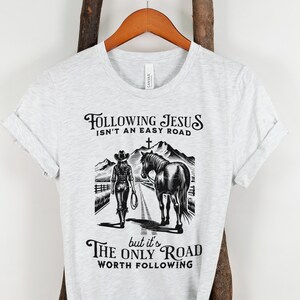 May include: A gray t-shirt with a black graphic design of a woman on horseback. The text reads "Following Jesus isn't an easy road, but it's the only road worth following."