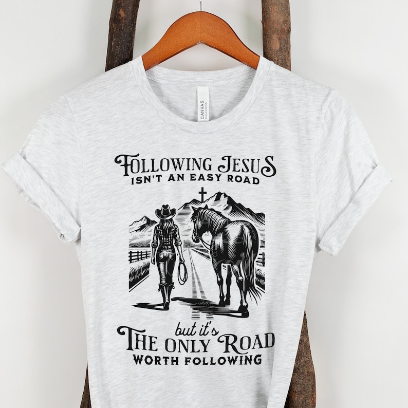 May include: A gray t-shirt with a black graphic design of a woman on horseback. The text reads "Following Jesus isn't an easy road, but it's the only road worth following."