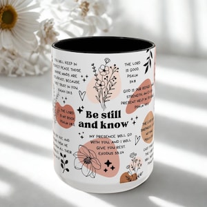 May include: White ceramic mug with a black interior, featuring the text "Be still and know" and various Bible verses. The design includes floral illustrations and pastel-colored shapes. A decorative and inspirational item.