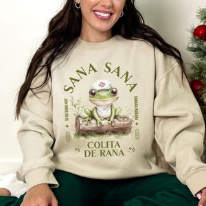 May include: A beige sweatshirt with a green frog wearing a nurse's hat and the text "Sana Sana Colita de Rana" in a circle around the frog. The text "Si no sana hoy, sanara ma&ntilde;ana" is written in a smaller font below the frog.
