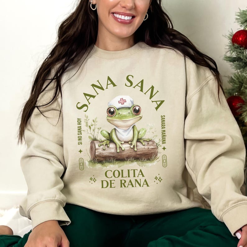 May include: A beige sweatshirt with a green frog wearing a nurse's hat and the text "Sana Sana Colita de Rana" in a circle around the frog. The text "Si no sana hoy, sanara ma&ntilde;ana" is written in a smaller font below the frog.