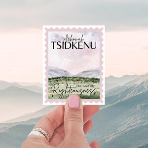 May include: A rectangular sticker with a pink border is held up against a mountain backdrop. The sticker features the words "Jehovah Tsidkenu" and "Righteousness" with a watercolor landscape scene.