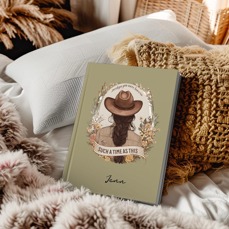 May include: A sage green journal with a decorative illustration of a woman wearing a cowboy hat, surrounded by flowers. The text reads "perhaps you were born for SUCH A TIME AS THIS" and "Jenn". The journal is resting on a bed with pillows and blankets.