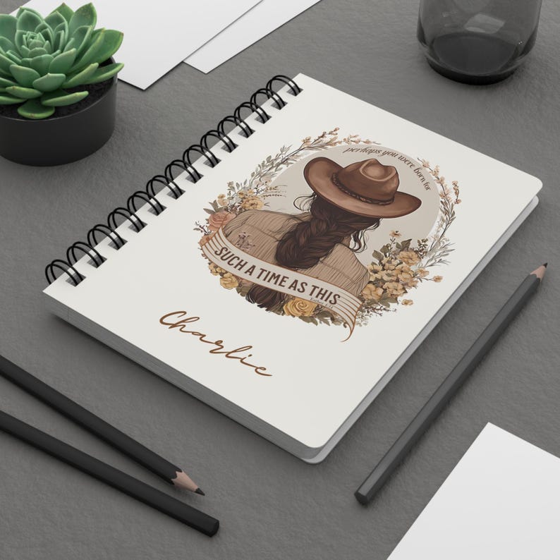 May include: A spiral-bound notebook with a cream-colored cover featuring an illustration of a woman wearing a cowboy hat. The illustration is surrounded by floral elements and the text "SUCH A TIME AS THIS" is displayed on a banner. The name "Charlie" is written below.