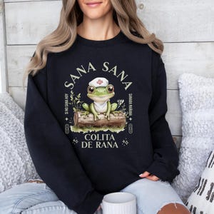May include: Black sweatshirt with a green frog wearing a nurse's hat. The frog is sitting on a log with green leaves. The text on the sweatshirt says "Sana Sana", "Sino Sana Hoy", "Samara Mariana", "Colita de Rana".