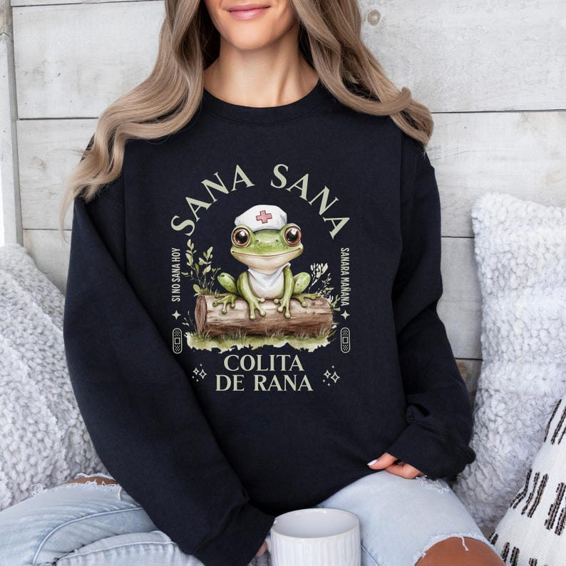 May include: Black sweatshirt with a green frog wearing a nurse's hat. The frog is sitting on a log with green leaves. The text on the sweatshirt says "Sana Sana", "Sino Sana Hoy", "Samara Mariana", "Colita de Rana".