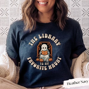 May include: Heather navy t-shirt with a Halloween-themed graphic. The design features a cartoon ghost reading a book inside an orange frame, surrounded by pumpkins. The text reads "THE LIBRARY IS MY FAVORITE HAUNT."