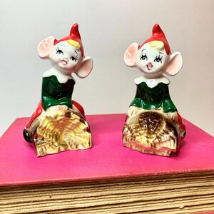 May include: A pair of vintage ceramic elf salt and pepper shakers. The elves are wearing green and red outfits and are sitting on a brown base.
