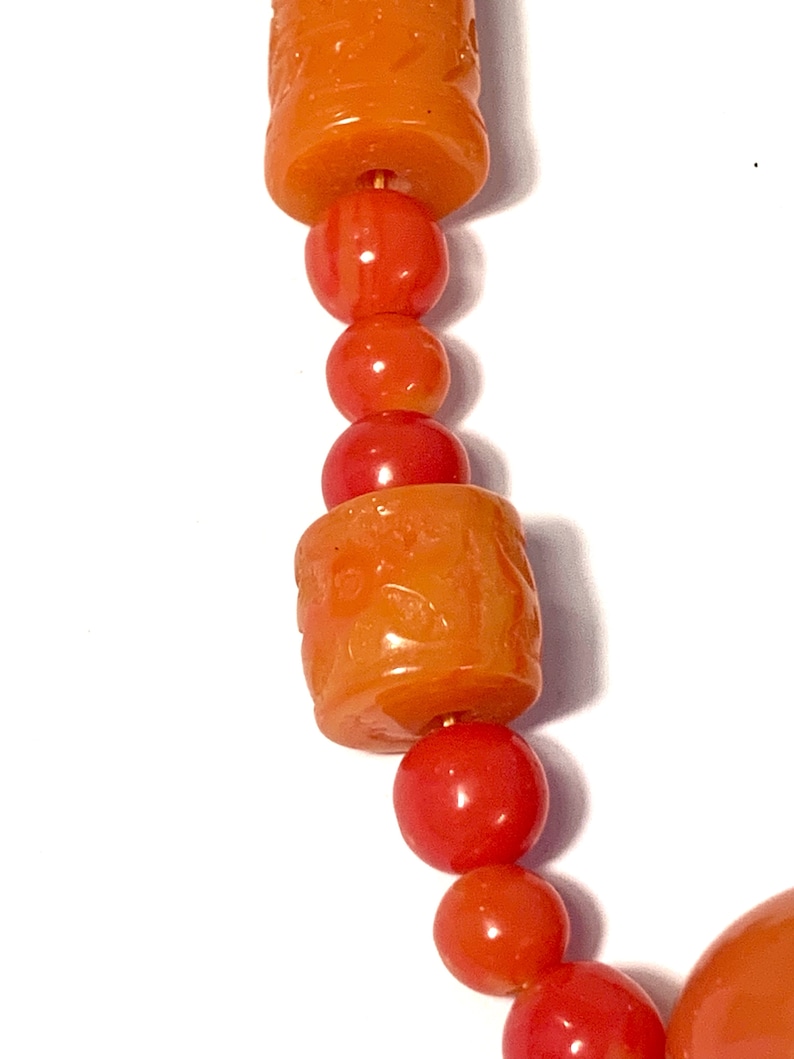 May include: A close-up of a necklace made of orange and red beads. The beads are round and have a smooth, polished finish. The necklace is strung on a thin wire.