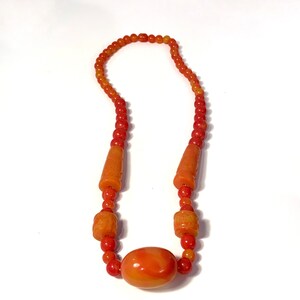 May include: A necklace made of orange and red beads with a large, oval-shaped bead in the center.