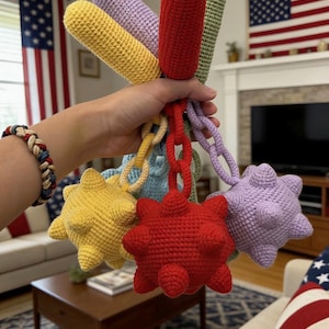 May include: A collection of colorful, crocheted toys in various shapes, including cylinders and spiky balls, connected by crocheted chains. The toys are in shades of red, yellow, purple, green, and blue. The background includes American flags and a living room setting.