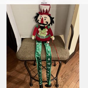 Puede incluir: Un muñeco cascanueces de peluche con chaqueta roja y detalles verdes, un sombrero alto rojo y blanco y pelo negro. El muñeco tiene largas piernas verdes de tela que cuelgan, cada una estampada con números blancos del uno al veinticuatro.
