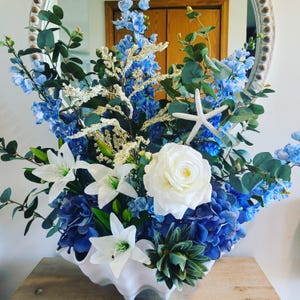May include: A floral arrangement featuring blue hydrangeas, delphiniums, and white lilies, accented with a white starfish and a large white rose. The flowers are arranged in a white, shell-shaped vase, creating a coastal theme.