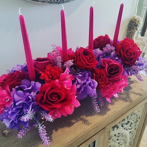 May include: A floral centerpiece with red roses, pink and purple flowers, and four tall pink candles. The arrangement sits on a wooden cabinet with carved details. The flowers include hydrangeas, carnations, and lavender.