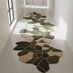 May include: A long, abstract rug with a geometric design in shades of green, black, and beige. The rug features irregular shapes with gold-colored lines separating the sections. It is placed in a bright hallway with a window.