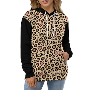 May include: A hooded sweatshirt with solid black sleeves and a tan leopard print body featuring repeating black cursive text that reads Jesus and small pink heart icons. The garment includes a front kangaroo pocket and white drawstrings.