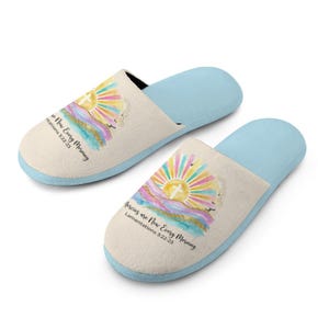 Puede incluir: Un par de zapatillas beige y azul claro. Cada zapatilla presenta un diseño de acuarela de un amanecer con una cruz y el texto "Mercies are New Every Morning, Lamentations 3:22-23". Las zapatillas tienen una textura suave y afelpada.